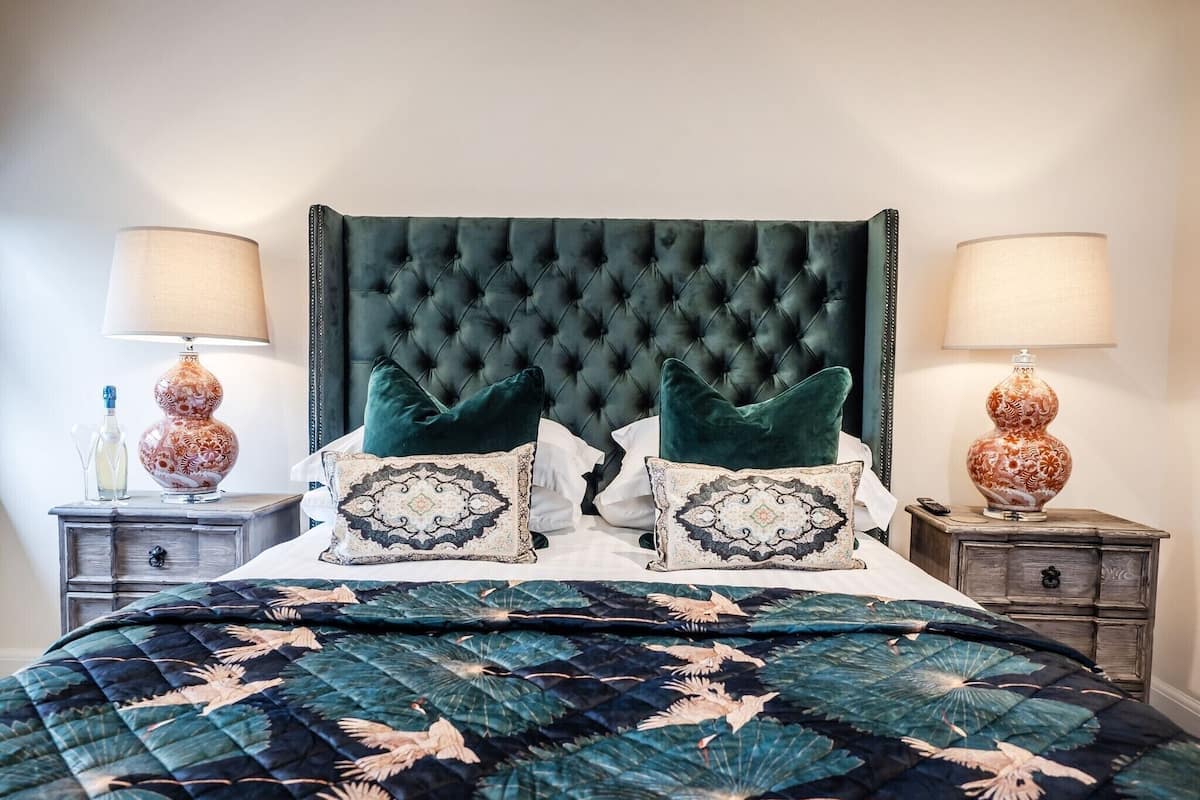 A bedroom with a tufted headboard, two bedside tables with lamps, a bed with a floral-patterned duvet, and decorative pillows.