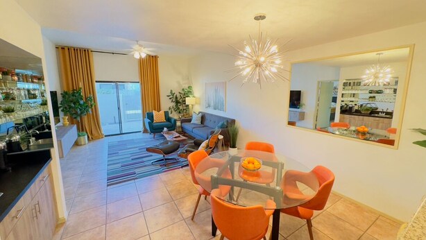 A modern living room with a glass dining table, orange chairs, and a sofa. There is a kitchen area with a countertop and a refrigerator.