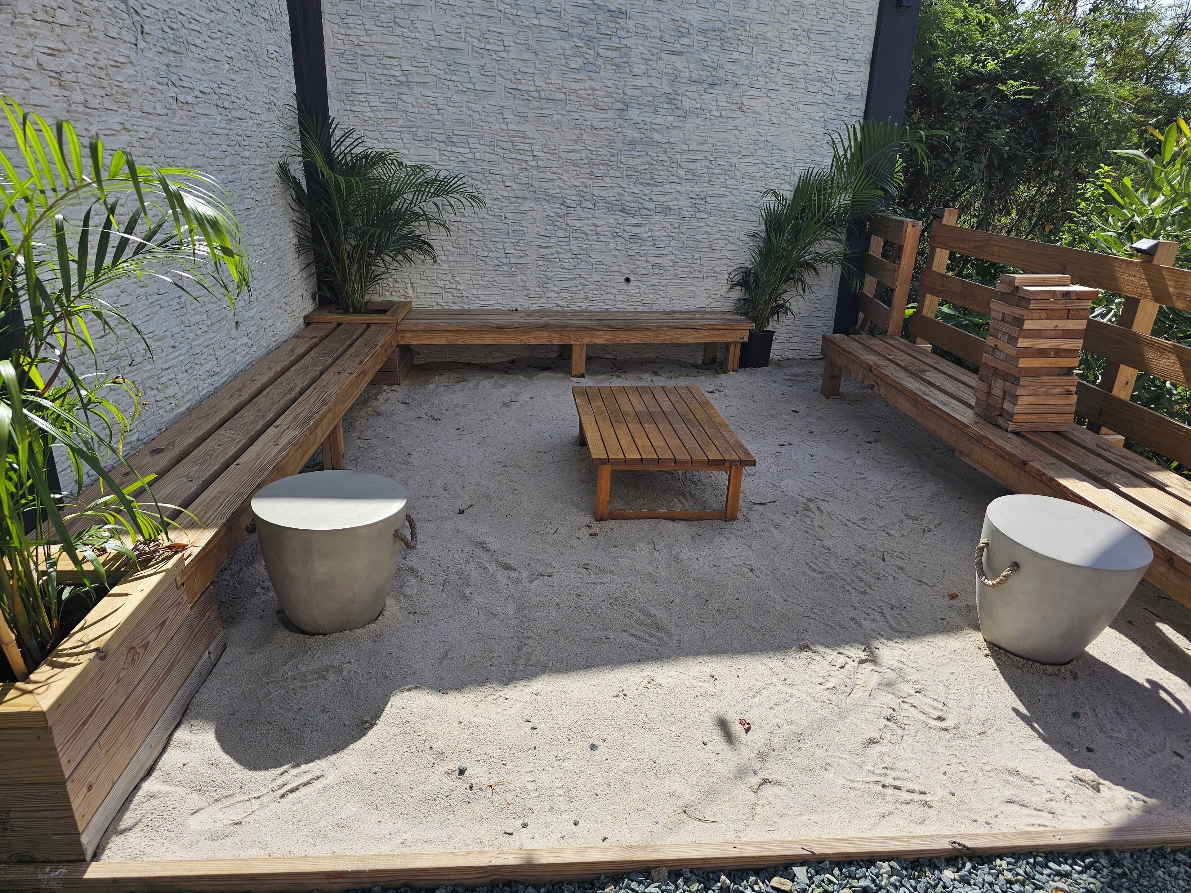 A sunlit outdoor seating area with wooden benches and tables, surrounded by sand and potted plants.