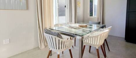 A dining area with a glass table and white chairs, a chandelier, and a painting on the wall.