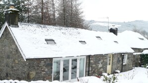 A stone house with a sloped roof covered in snow, a chimney, and a door leading to a glass door. - Burn Cottage - A Secluded Highland Retreat For Two (Fortingall)