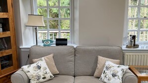A living room with a grey sofa, two floral cushions, a wooden side table, a lamp, and a bookshelf. - Burn Cottage - A Secluded Highland Retreat For Two (Fortingall)