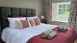 A neatly made bed with a red blanket and rolled towels, a green upholstered headboard, and a window with floral curtains. - Burn Cottage - A Secluded Highland Retreat For Two (Fortingall)