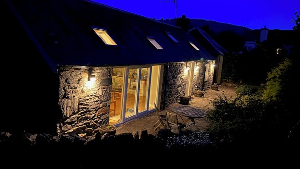 A stone house with a sloped roof and skylights, featuring a glass door entrance and a small patio with outdoor seating. - Burn Cottage - A Secluded Highland Retreat For Two (Fortingall)