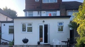 Una casa de dos pisos con puerta principal, un pequeño porche y un patio con parrilla y sillas.