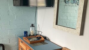 Ein Zimmer mit an der Wand montiertem Fernseher, einem Holzkasten mit Schubladen, einem Korbstuhl und einem gerahmten Kunstwerk an der Wand.