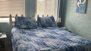 Ein Schlafzimmer mit einem großen Bett, zwei Fenstern mit Jalousien, einem Wandbild und einem kleinen Nachttisch.