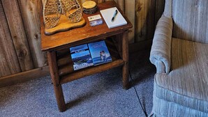 A wooden wall with a framed picture of birds, a lamp, a notepad, and a small wooden table with a woven basket and brochures. - Cozy Beachcomber Cottage on Great Lake Huron (Ausable Township)