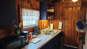 A compact kitchen with wooden walls, a window with a white valance, a microwave, a coffee maker, and a sink. - Cozy Beachcomber Cottage on Great Lake Huron (Ausable Township)