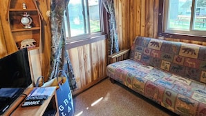 A cozy cabin room with wooden walls, a sofa with a wildlife-themed cover, a wooden shelf with a clock, and a window with curtains. - Cozy Beachcomber Cottage on Great Lake Huron (Ausable Township)