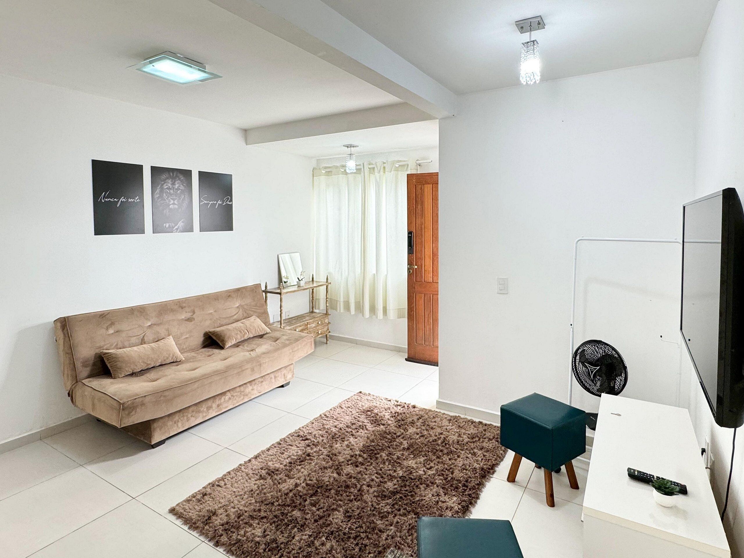 Ein Wohnzimmer mit einem beigefarbenen Sofa, einem braunen Shaggy-Teppich, einem weißen Couchtisch, einem türkisfarbenen Hocker, einem Fernseher und einem Ventilator.