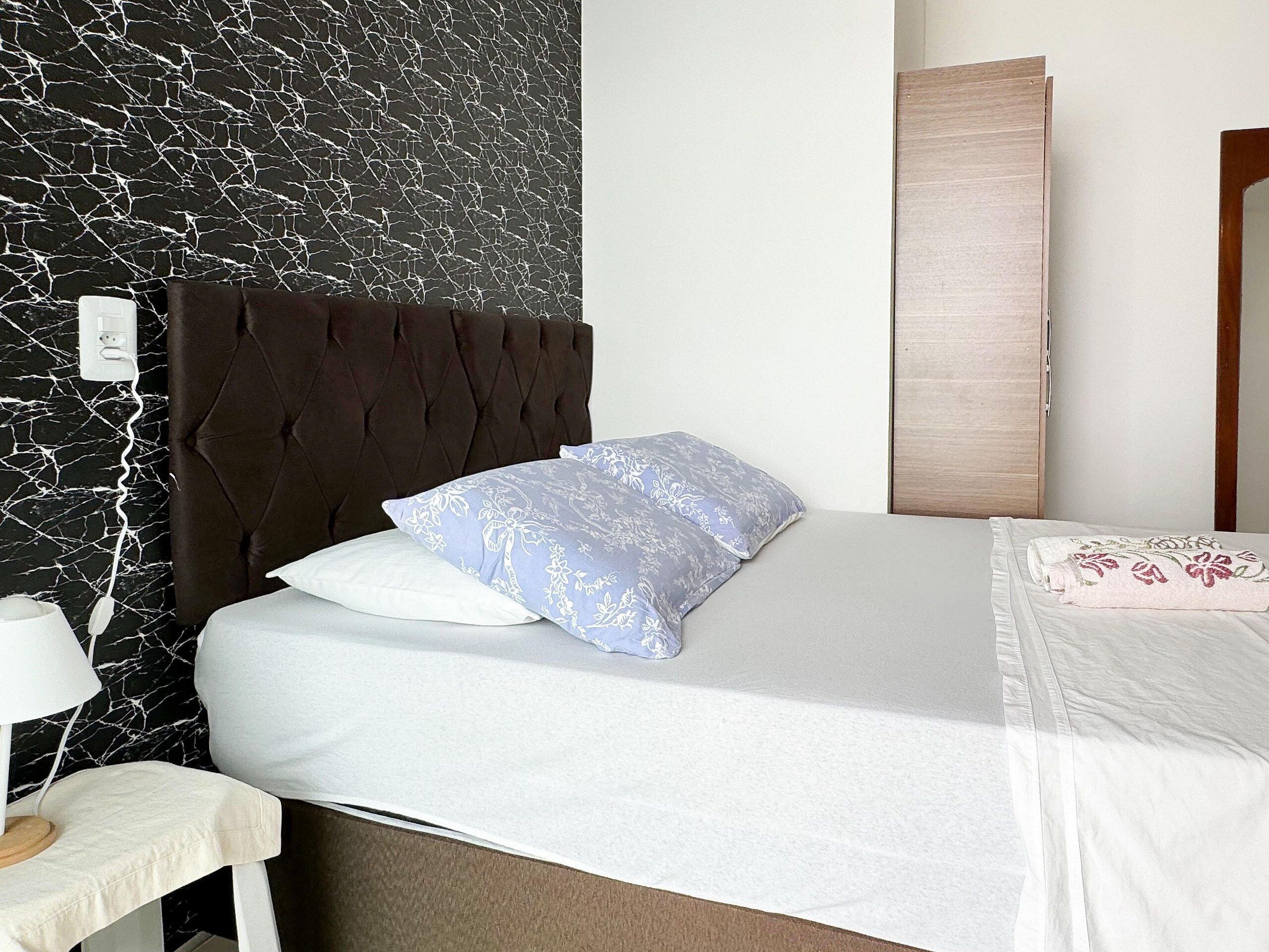 A neatly made bed with a white and light blue floral pillow, a dark brown upholstered headboard, and a white nightstand with a lamp.