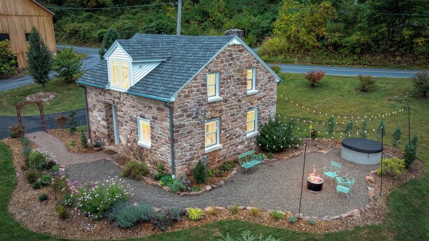 Une maison en pierre avec un toit en pente, un foyer extérieur, un espace salon de jardin et un trampoline dans la cour.