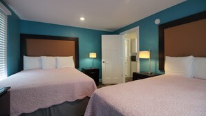 Habitación de hotel con dos camas, una puerta que da a un baño y una mesita de noche con lámpara.