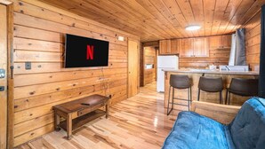 A cozy log cabin interior with a wooden TV mounted on the wall, a small wooden table, a blue sofa, and a kitchen area with a refrigerator and bar stools.