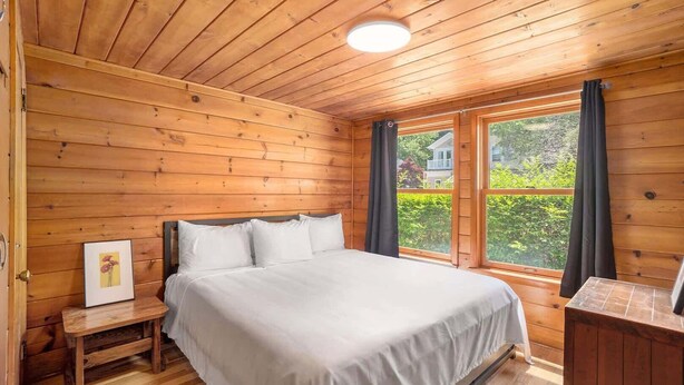 A wooden cabin bedroom with a bed, a nightstand, a framed picture, and a window with curtains.