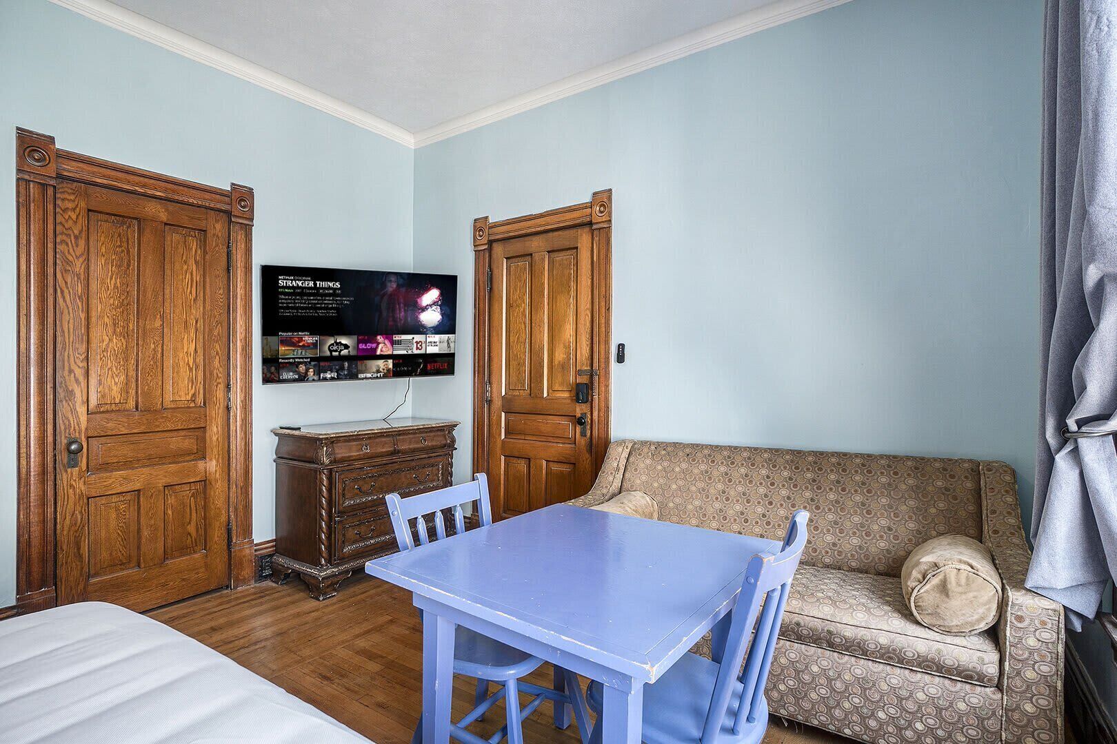 Une pièce comprenant une armoire en bois, un téléviseur fixé au mur, une table bleue avec des chaises et un canapé à motifs.