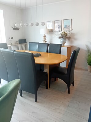 Uma área de refeições com uma mesa redonda, seis cadeiras, luminárias pendentes, quadros, um radiador e um vaso de planta.