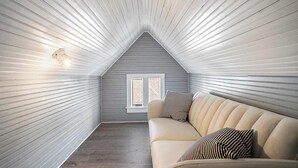 Un dormitorio abrigado en el ático, con techo inclinado, un sofá color beige y almohadas a rayas, y una ventana con contraventanas blancas.