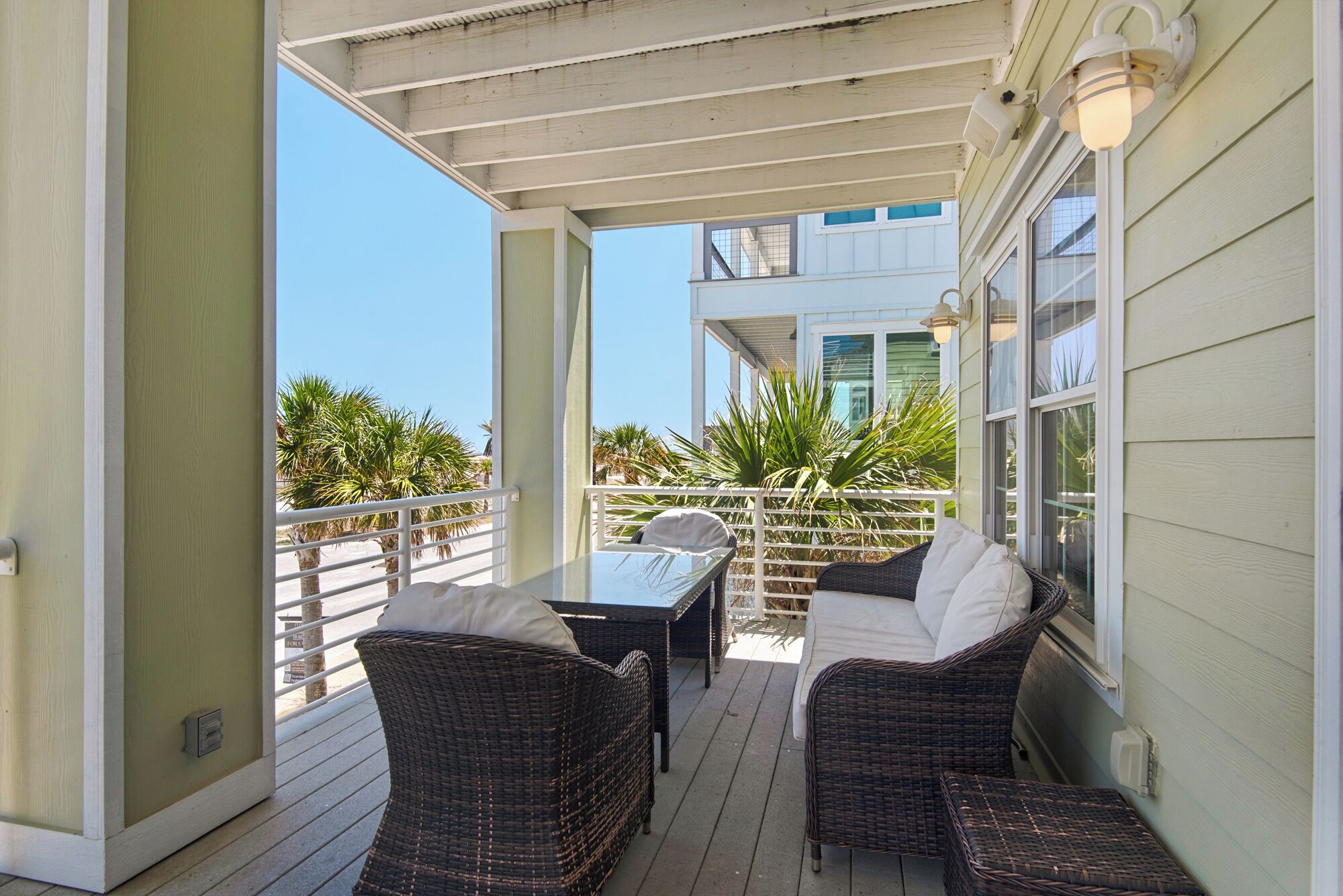 A covered outdoor patio with wicker furniture and a table, overlooking a building with a balcony.