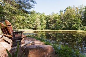 Ein Holz-Adirondack-Stuhl an einem Teich, umgeben von dichtem Wald.
