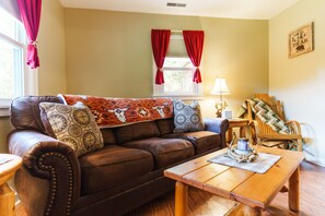 A cozy living room with a brown leather sofa, a wooden coffee table, and a window with red curtains.