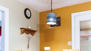 A dining area with a round wooden table and two wooden chairs, a hanging light fixture, a wall clock, and a small shelf with a decorative item. - Woodland view-Cedar Cottage (Laurelville)