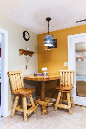 Un coin repas comprenant une table ronde en bois et deux chaises en bois, un luminaire suspendu, une horloge murale et une petite étagère sur laquelle est posé un objet décoratif.