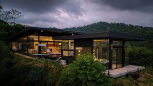 Ett modernt hus med en swimmingpool, stora glasfönster och utsikt över en skogbeklädd kulle.