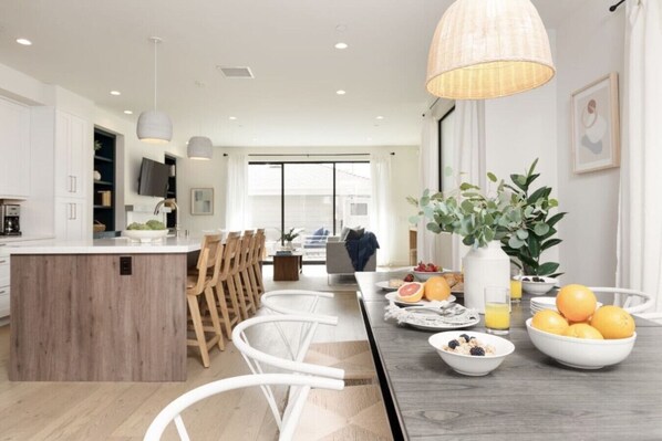 A modern kitchen with a central island, wooden chairs, and a dining table set with fruits and breakfast items.