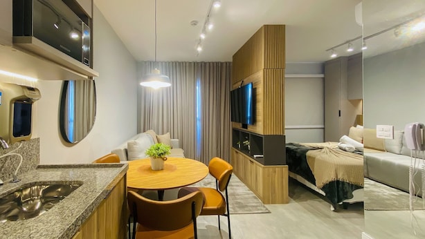 A modern living room with a dining area, a TV mounted on a wooden panel, and a built-in kitchen with a granite countertop. - Essence Studio Vila Mariana | Air Conditioning, Wi-Fi & Vila Mariana Metro (São Paulo)