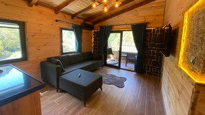 A cozy log cabin interior with a sectional sofa, a small ottoman, a stone fireplace, and a balcony with a stone railing. - Nika Otel (Camlihemsin)