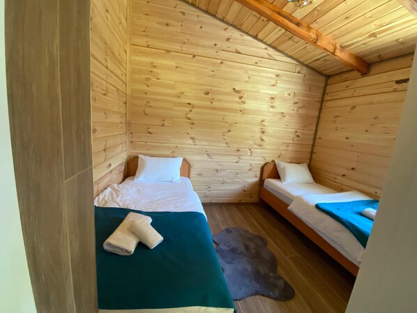 Una habitación pequeña y acogedora con paredes de madera y techo inclinado. Hay dos camas, con ropa blanca y una manta verde oscuro en una de ellas. Un tapete pequeño y oscuro cubre el suelo de madera.