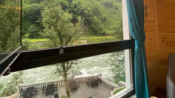 A room with a large window overlooking a river and a forested hillside. - Nika Otel (Camlihemsin)