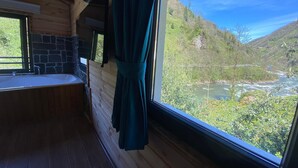 A wooden cabin interior with a stone wall, a sink, and a window overlooking a river and forested hills. - Nika Otel (Camlihemsin)