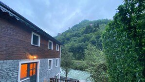 A building with a stone facade and wooden upper levels, outdoor seating area with tables and chairs, and a view of a river and forested hills in the background. - Nika Otel (Camlihemsin)
