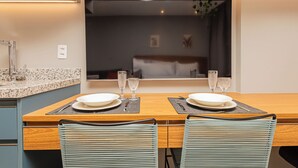 Un comedor moderno con una mesa de madera dispuesta para dos personas, dos sillas y vistas a un dormitorio a través de un ventanal grande.