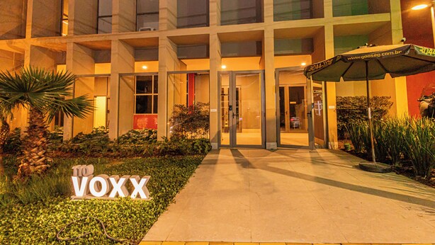 Un edificio moderno con entrada de vidrio, un letrero que dice "VOXX" y un área exterior bien iluminada con plantas y un paraguas.