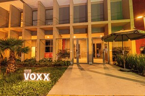Un edificio moderno con entrada de vidrio, un letrero que dice "VOXX" y un área exterior bien iluminada con plantas y un paraguas.