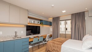 Una habitación de hotel moderna con kitchenette, una cama, un escritorio con silla y un ventanal con cortinas.