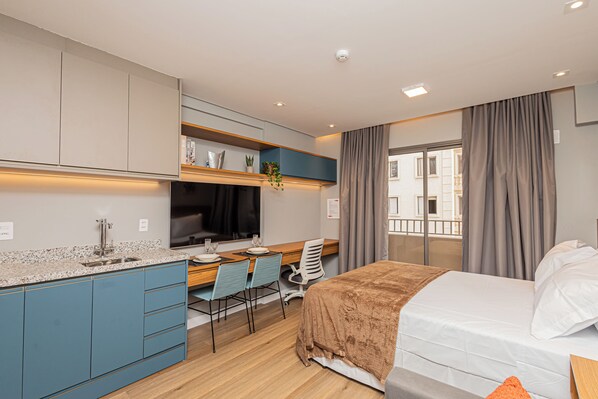 Una habitación de hotel moderna con kitchenette, una cama, un escritorio con silla y un ventanal con cortinas.