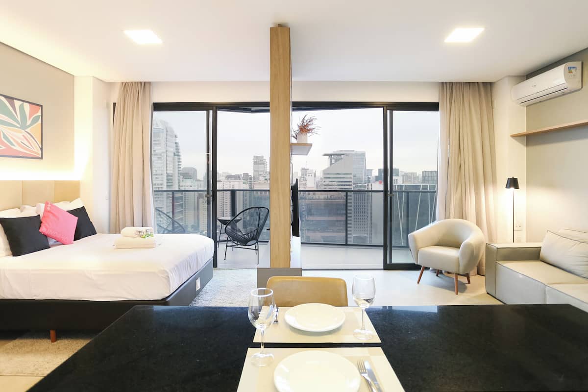 Ein Hotelzimmer mit einem Bett, einem Esstisch gedeckt mit Tellern und Gläsern, einem Sofa, einem Sessel und einem Balkon mit Blick auf die Stadt.