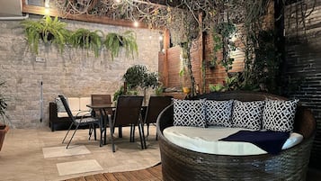 Uma área externa com assento, incluindo um sofá de vime redondo, uma mesa de jantar com cadeiras e plantas em vasos.