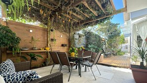 Un patio exterior con mobiliario de mimbre, una pérgola de madera y diversas macetas con plantas.