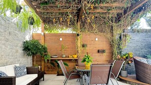 Un patio exterior con una pérgola de madera, muebles de mimbre y plantas en macetas.