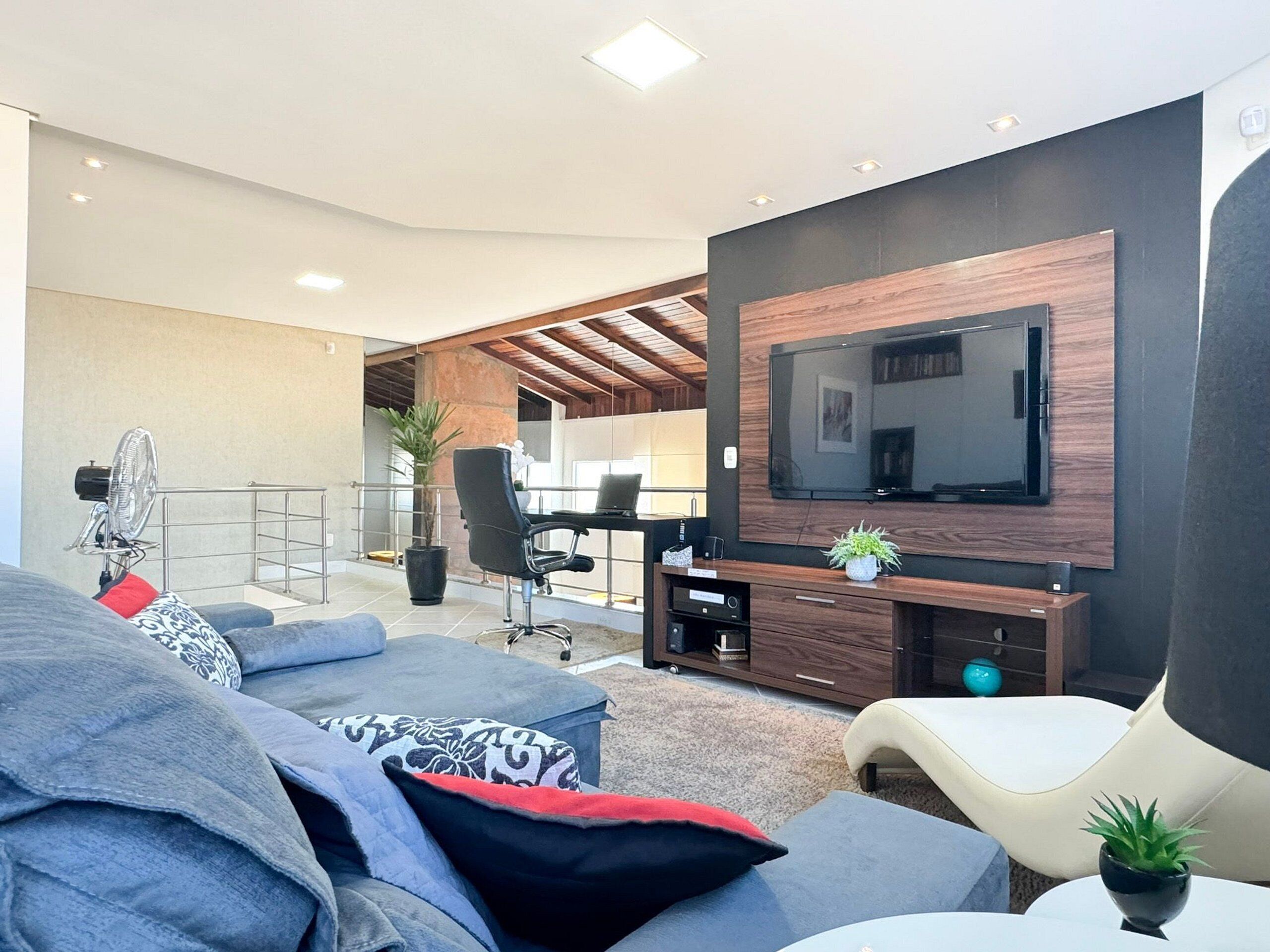 Uma sala de estar moderna com uma grande TV de tela plana presa em uma parede escura, um sofá confortável com almofadas estampadas, uma pequena mesa redonda e um espaço de escritório com uma cadeira e um vaso de planta.