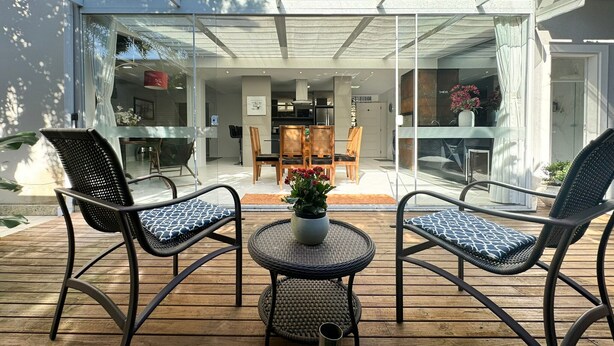 Un patio al aire libre con dos sillas de mimbre y una pequeña mesa, un área de comedor con paredes de vidrio y vistas del interior.