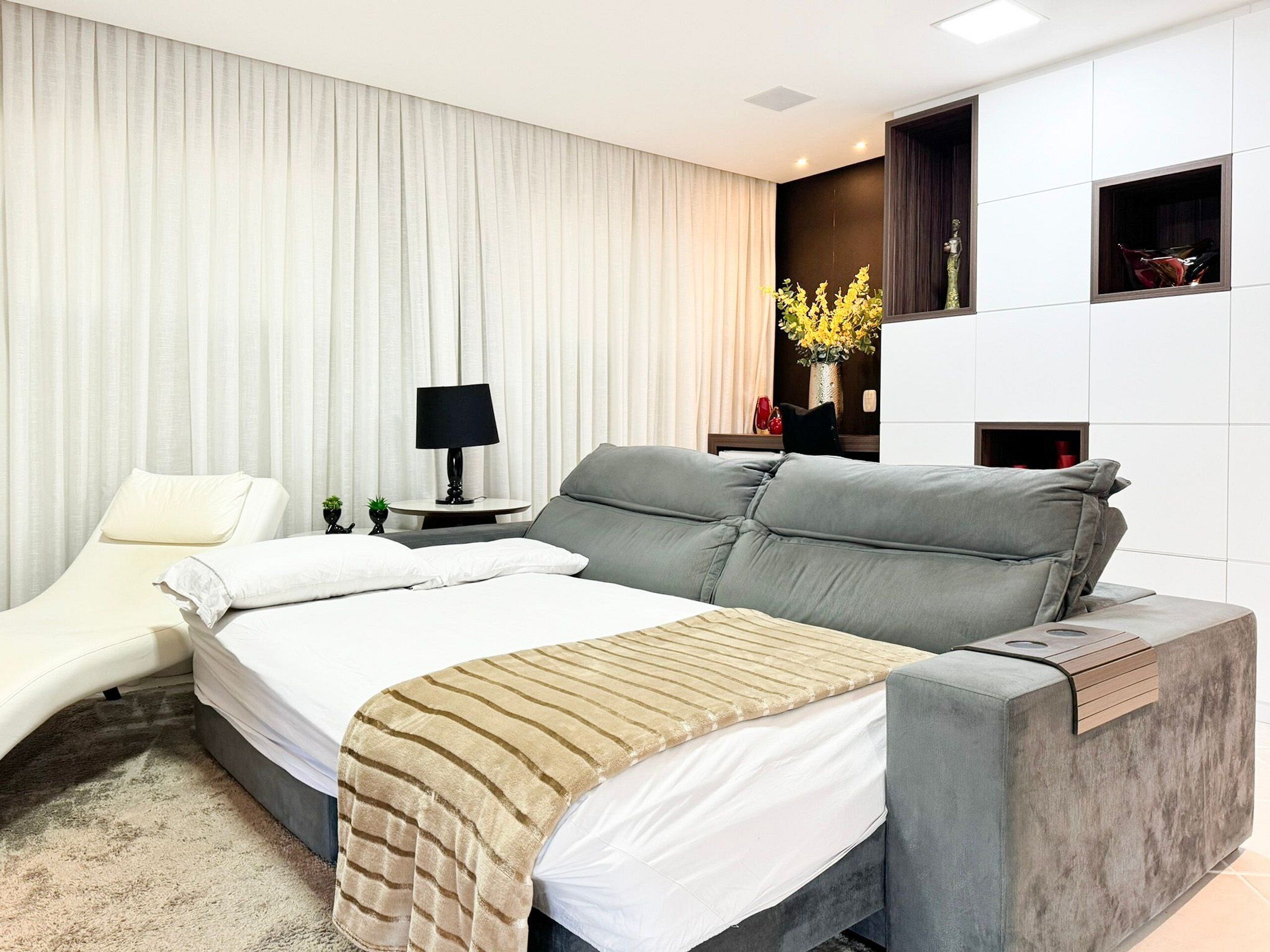 A modern hotel room with a large bed, a bedside table with a lamp, a small plant, and a decorative vase with flowers.