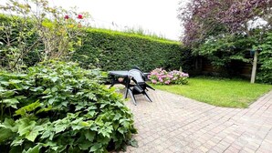 Ein gepflasterter Gartenweg mit einem schwarzen Metalltisch und -stühlen, umgeben von üppiger Bepflanzung und blühenden Pflanzen.