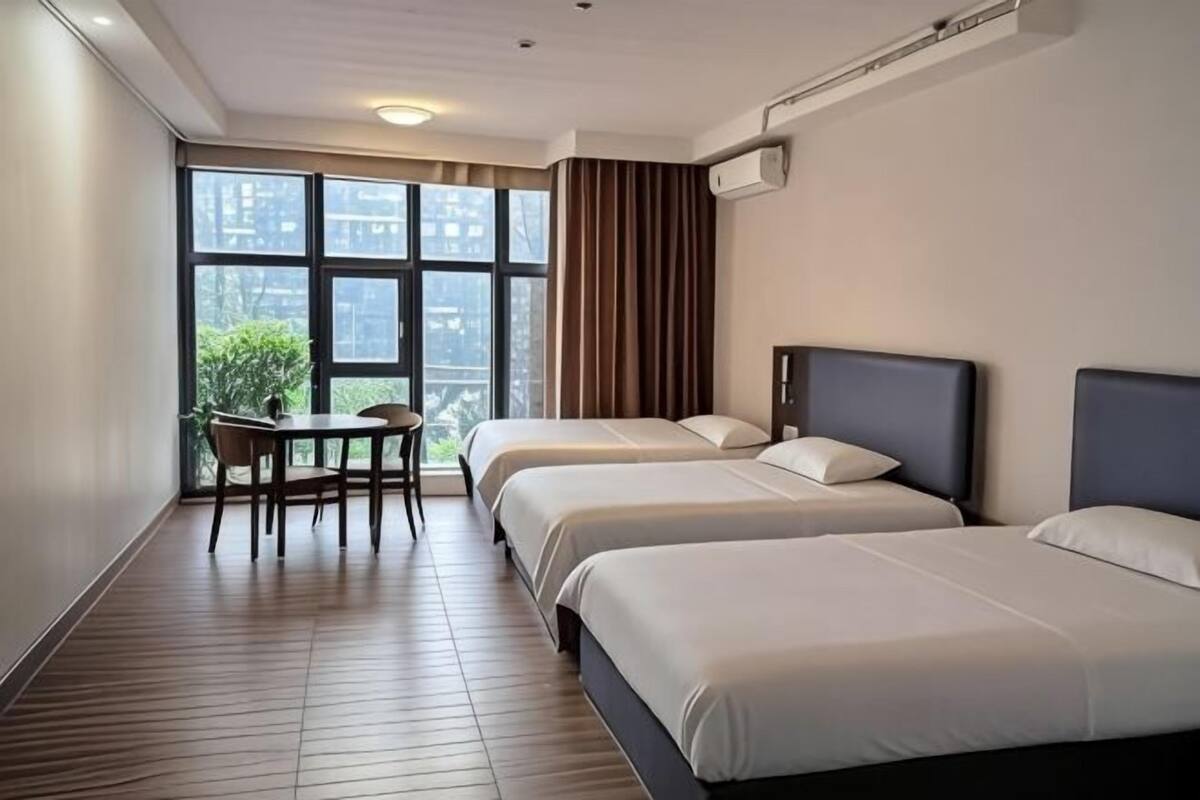 Une chambre d’hôtel avec trois lits, une petite table avec une chaise et une grande fenêtre avec des rideaux.